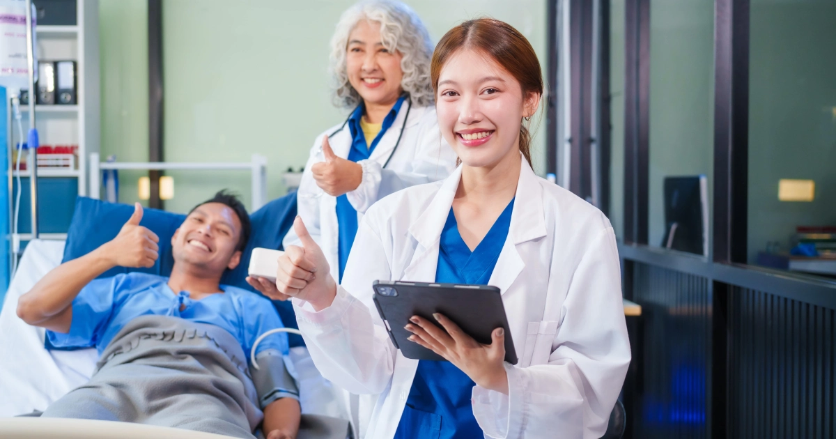 Two female medical professionals in a hospital room giving thumbs-up, one holding a tablet, with a smiling patient enjoying painless treatments in Dallas.